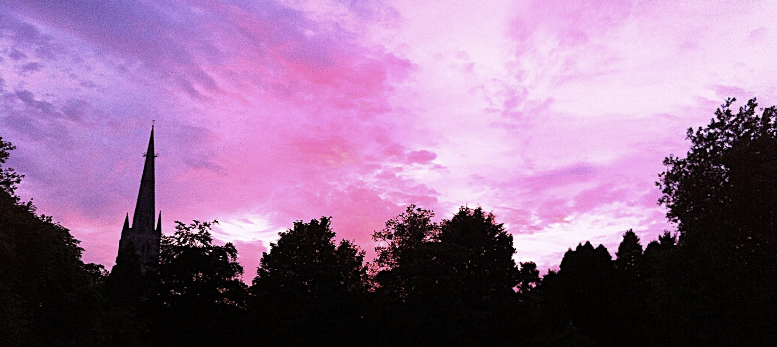 A silhouette of a church spire and some trees. The sky is purple, pink and blue with a beautiful sunset